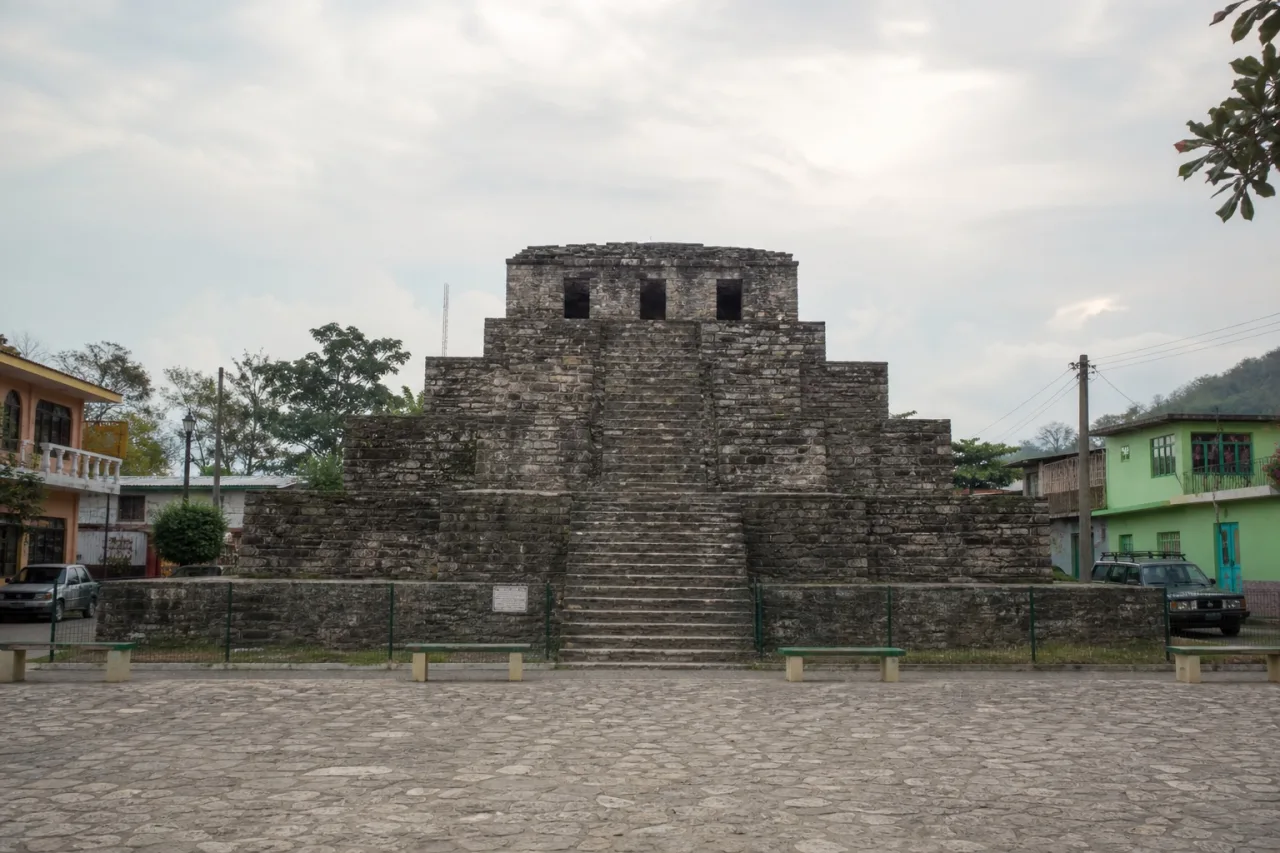 Pirámide de piedra con escalinata central y templo en la cima, en una plaza de pueblo con casas de color.